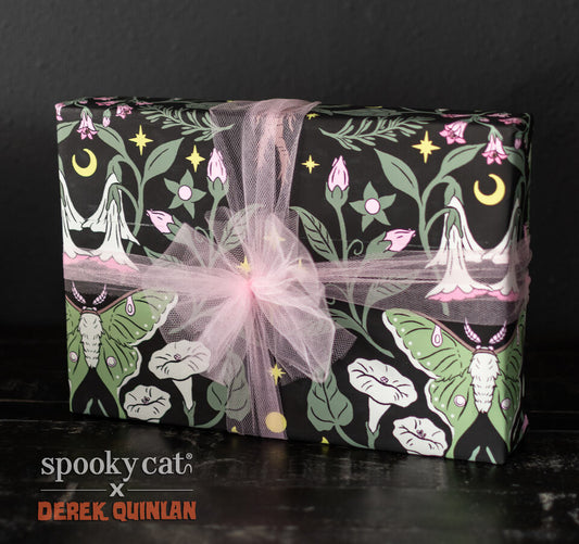 Gift wrapped in Mandrake Floral wrapping paper featuring green Luna Moths, pink belladonna flowers, crescent moons, stars, and small mandrake root figures on black background, tied with pink tulle ribbon.
