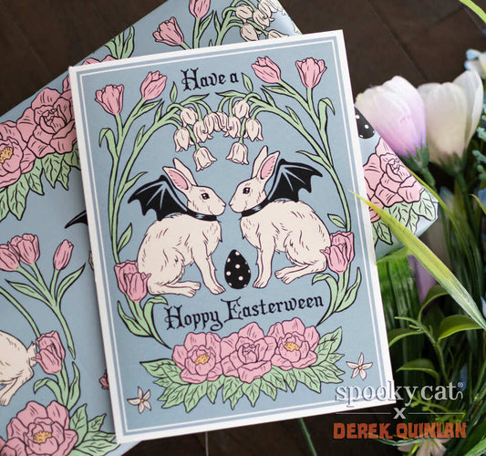 Greeting card reading “Have a Hoppy Easterween” featuring two white Vampire Bunnies with bat wings, surrounded by pink flowers and a black striped egg on blue background, displayed over matching wrapping paper beside spring flowers.