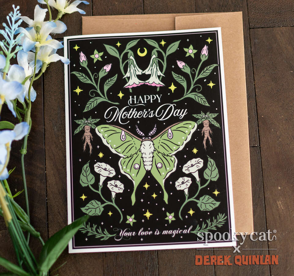 Luna moth Mother’s Day card with green moth, mandrake roots, and gothic floral design on black background, displayed on wood with white flowers