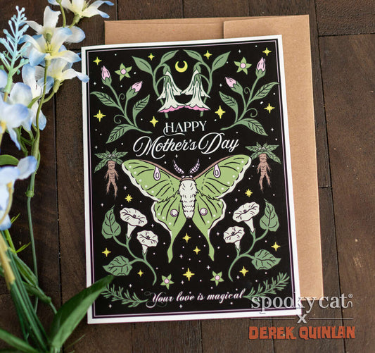Luna moth Mother’s Day card with green moth, mandrake roots, and gothic floral design on black background, displayed on wood with white flowers