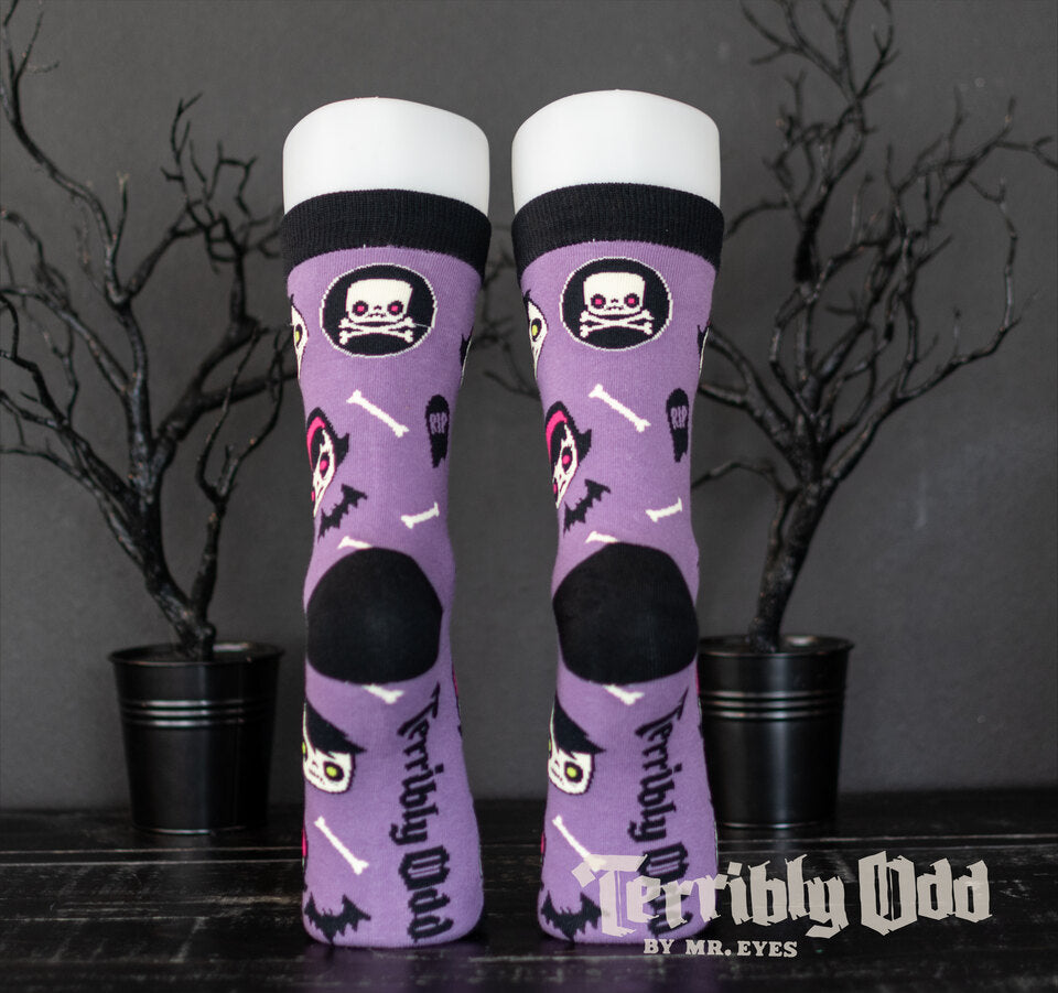 Terribly Odd Coffin Kid socks featuring a boy and girl, bats, bones, and tombstones on a purple background worn on a foot mannequin.