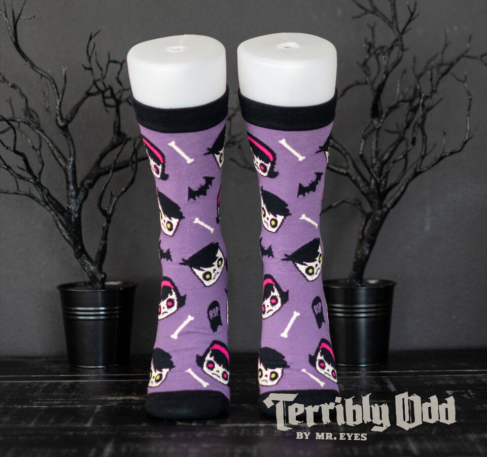 Terribly Odd Coffin Kid socks featuring a boy and girl, bats, bones, and tombstones on a purple background worn on a foot mannequin.