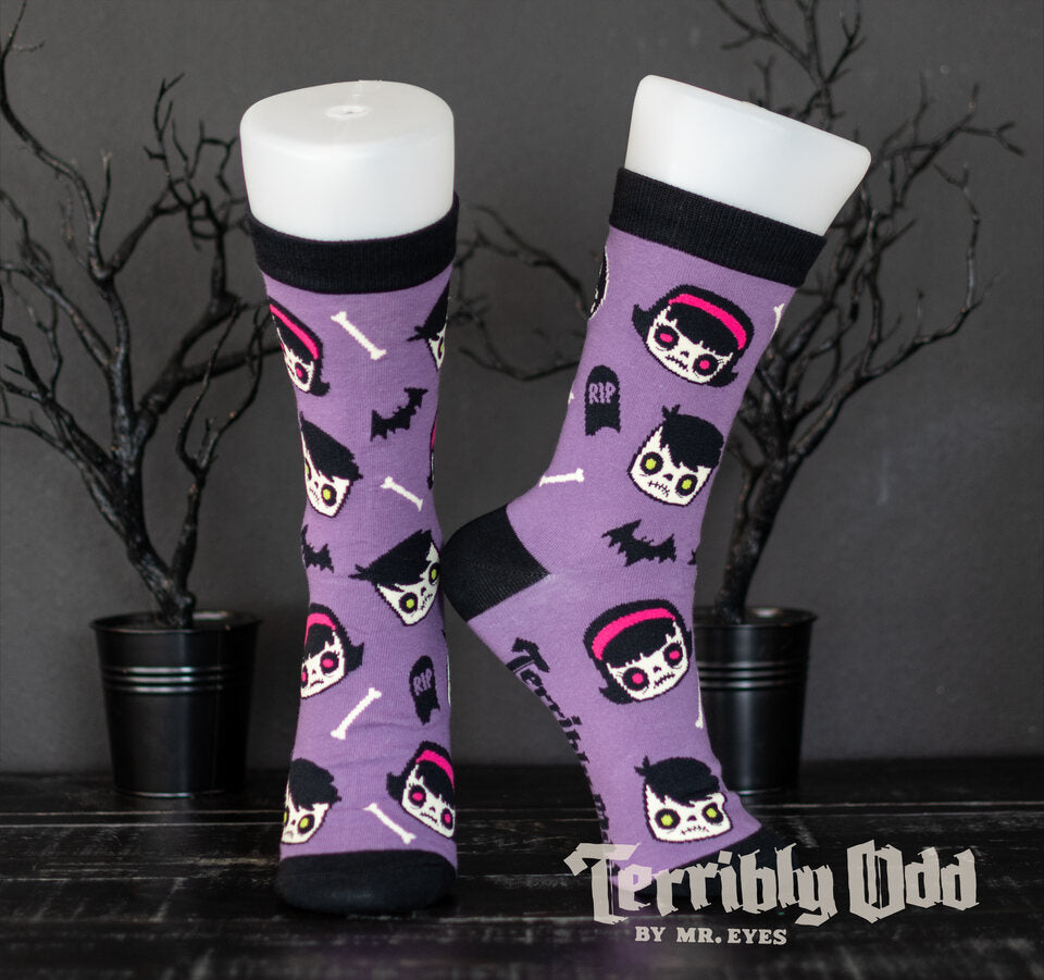 Terribly Odd Coffin Kid socks featuring a boy and girl, bats, bones, and tombstones on a purple background worn on a foot mannequin.
