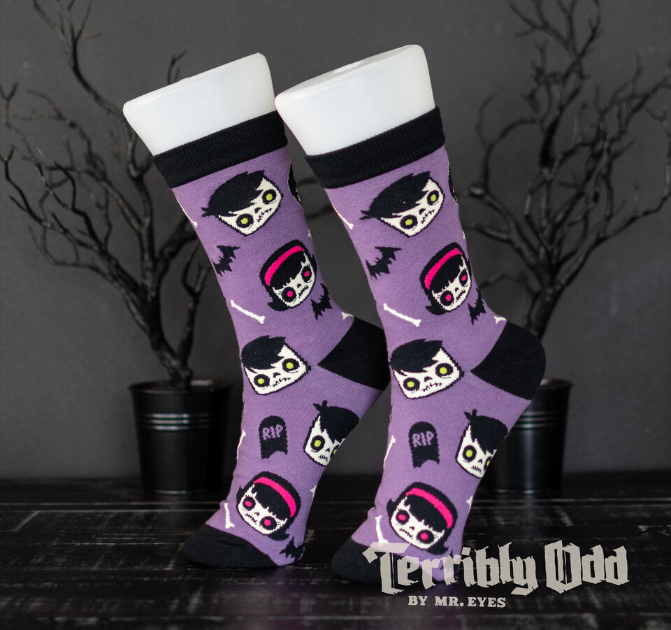 Terribly Odd Coffin Kid socks featuring a boy and girl, bats, bones, and tombstones on a purple background worn on a foot mannequin.