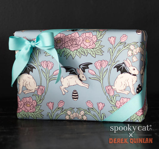 Wrapped gift in light blue Vintage Easterween wrapping paper featuring Vampire Bunnies with bat wings, pink flowers, and striped black Easter eggs, tied with a teal ribbon bow.