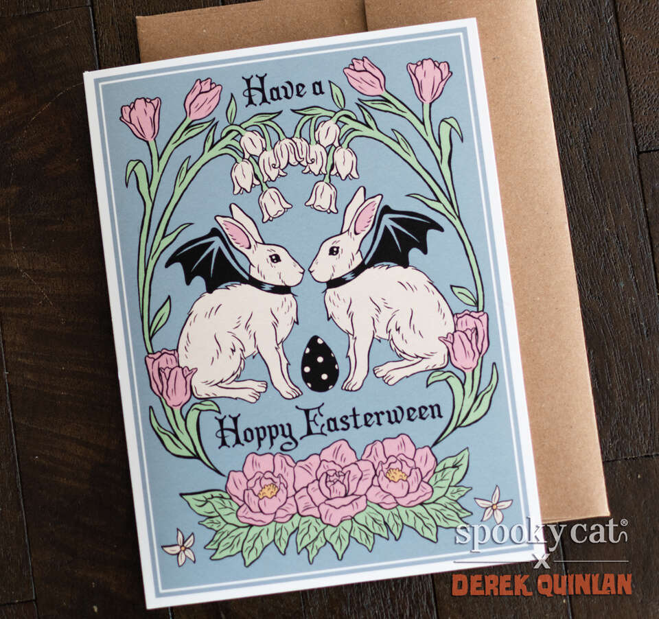 Vintage Easterween greeting card with two bat-winged bunnies and pink floral border on blue background, shown with kraft envelope on dark wooden surface.