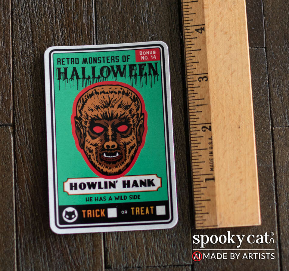 A rectangular Halloween sticker featuring a vintage-style illustrated monster head with the text 'RETRO MONSTERS OF HALLOWEEN' and 'HOWLIN' HANK' is shown on a dark wood surface next to a wooden ruler for scale. 
