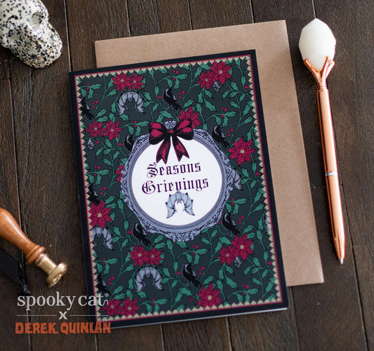 Creepmas greeting card reading “Seasons Grievings” in a round victorian frame surrounded by red poinsettias, green foliage, ravens, and bats, displayed in front of a kraft envelope on a dark wood background. Designed by Derek Quinlan for Spooky Cat Press.